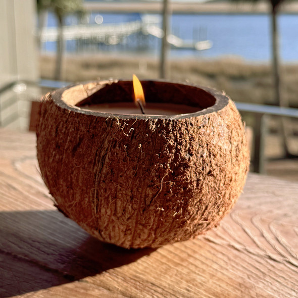 A unique, hand-poured candle in a natural, hollowed-out coconut shell is lit and glowing, sitting on an outdoor wooden table with a beautiful, sunny coastal scene in the background.