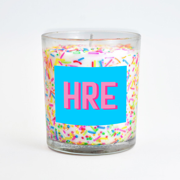 The festive sprinkles inclusion candle in a clear glass jar, shown with a personalized blue label that reads "HRE." The festive sprinkles inclusion candle in a clear glass jar, shown with a personalized blue label that reads "HRE."