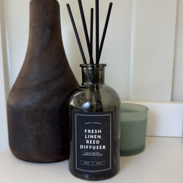 A Fresh Linen reed diffuser, featuring a dark gray glass bottle and black cotton reeds, is elegantly styled on a shelf next to a dark wooden vase and a candle, creating a sophisticated and calming home decor scene. A Fresh Linen reed diffuser, featuring a dark gray glass bottle and black cotton reeds, is elegantly styled on a shelf next to a dark wooden vase and a candle, creating a sophisticated and calming home decor scene.