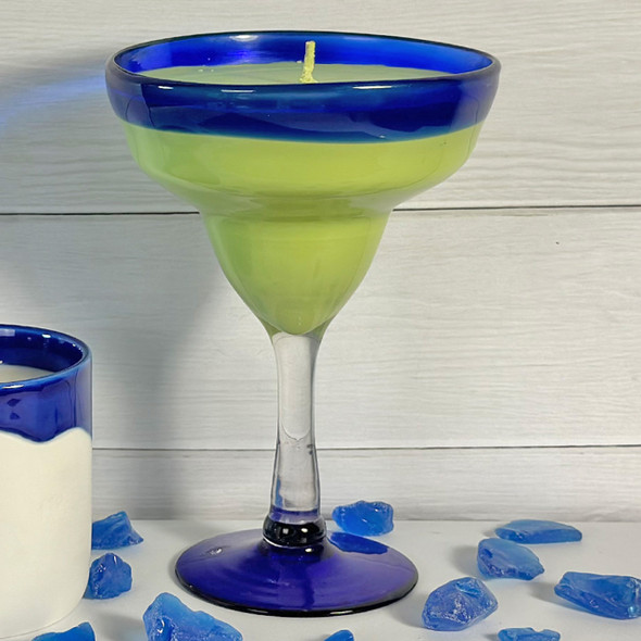 A festive La Playa Cocktail Candle, hand-poured into a blue and green margarita glass, is displayed on a surface with blue sea glass, evoking a tropical beach vibe.