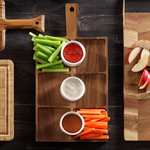 Overhead of acacia serving board with six compartments, celery and carrots, and three white ceramic ramekins of dip