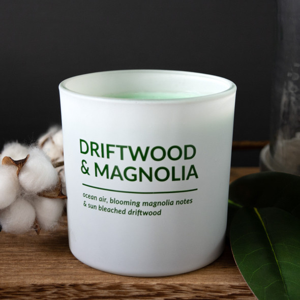 A Driftwood & Magnolia scented soy candle in a stylish, matte white printed glass jar is displayed on a wooden surface, elegantly styled with natural cotton bolls and a green leaf.