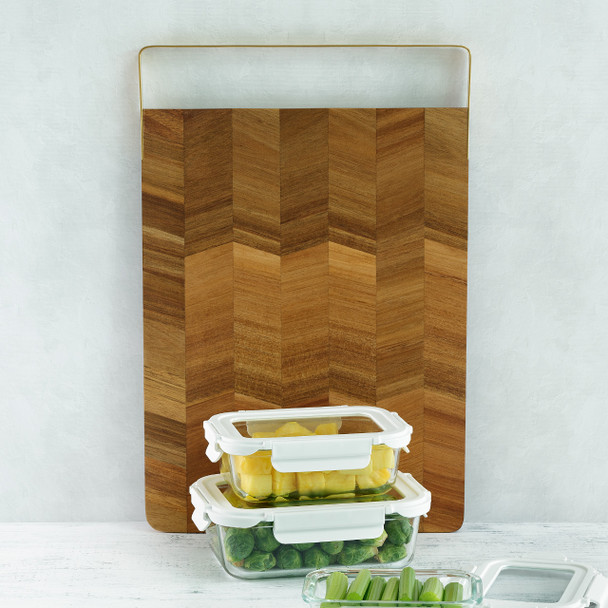 A rectangular acacia wood board with a striking end-grain chevron pattern and a modern matte gold handle is displayed vertically in a kitchen, with glass meal prep containers in front of it.
