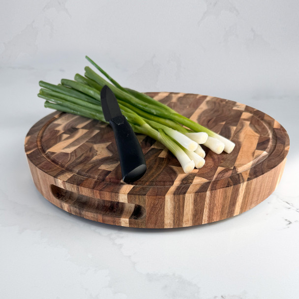 A beautiful Acacia Round End Grain Board is displayed on a white marble countertop, holding a fresh bunch of green onions and a small paring knife, ready for vegetable prep.