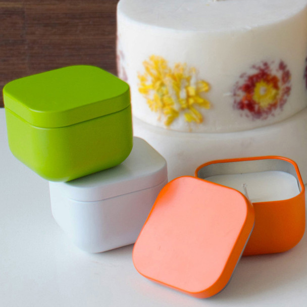 A set of three modern, square candle tins in lime green, white, and orange are artfully arranged, with the orange tin open to reveal the soy wax candle inside.