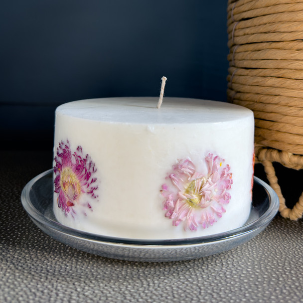 A large, white, hand-crafted inclusion candle, with real pink and red flowers embedded in the outer wax, is elegantly displayed on its included reusable glass tray against a dark, moody background.
