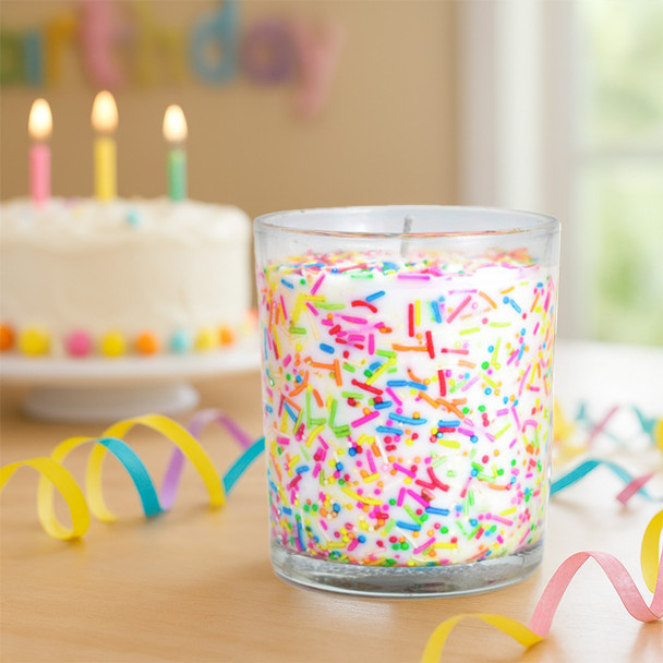 Bright sprinkle-filled candle in a clear glass jar, set on a wooden table surrounded by colorful ribbon streamers, with a white frosted birthday cake and lit candles softly blurred in the background.