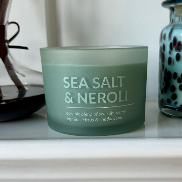 A Sea Salt & Neroli scented soy candle in an elegant, frosted sage green glass jar is styled on a white shelf next to decorative glassware, creating a serene, coastal-inspired scene.