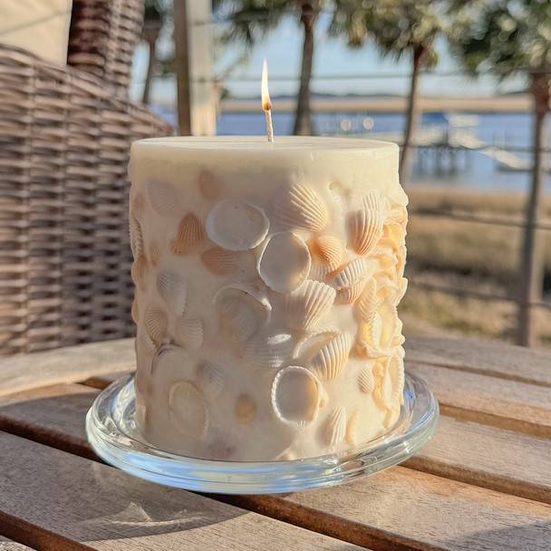 A beautiful, hand-crafted inclusion candle covered in real seashells is lit and glowing warmly, sitting on a glass tray on an outdoor table with a scenic coastal view in the background.