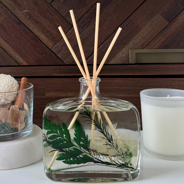 A decorative Dream Garden Inclusion Reed Diffuser in a clear glass bottle, featuring beautiful green botanical inclusions and natural wood reeds, is styled on a white surface next to a candle and other home decor.