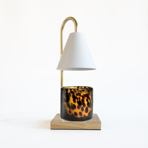 Front view of the candle warmer with jar candle beneath the shade on a warm wood base, studio backdrop