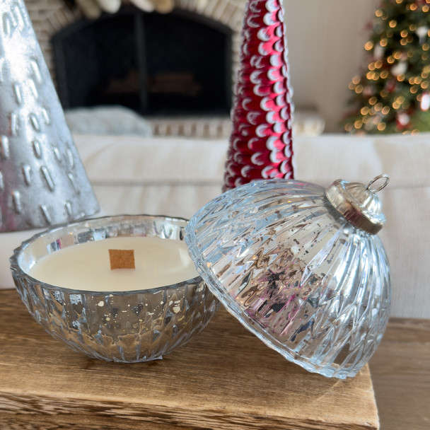A beautiful Silver Bell Ornament Candle made of ridged, silver mercury glass is displayed open on a wooden mantel, revealing the wood wick candle inside, with a cozy fireplace and Christmas tree in the background.