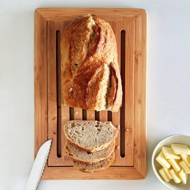Overhead shot of a bamboo bread board with removable slatted crumb tray holding a sliced artisan loaf, bread knife and a small bowl of butter on the counter.