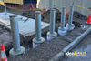 Steel safety bollards set in fresh concrete footings along a curbside construction area with dirt piles, cones, and caution fencing nearby