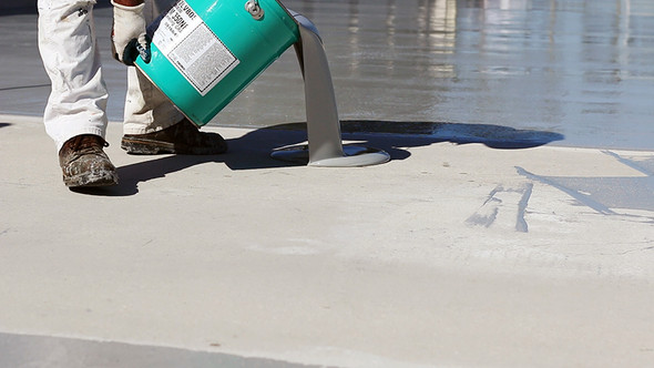 Contractor pouring gray polyurethane waterproofing coating from a teal pail onto a concrete surface.