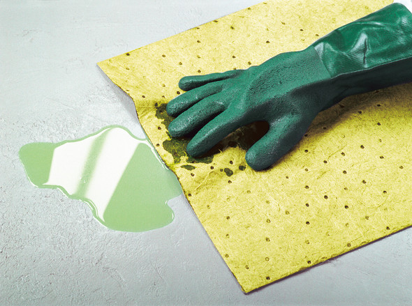 Hand in a green protective glove using a yellow chemical absorbent pad to soak up a hazardous liquid spill on a floor.