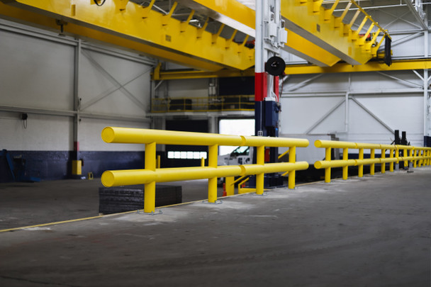 Long yellow steel guardrail system installed inside a large industrial warehouse, providing heavy-duty impact protection along equipment and traffic lane