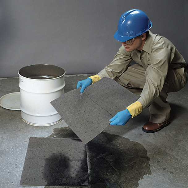 Worker wearing protective gear placing gray absorbent pads onto a liquid spill next to an industrial drum.