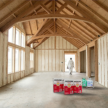 Interior of unfinished wood-framed room with spray foam insulation applied between studs and exposed vaulted ceiling beams; worker exiting in protective suit with foam kits placed on the floor.
