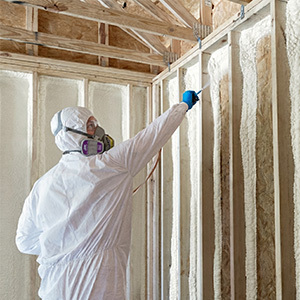 Interior of unfinished wood-framed room with spray foam insulation applied between studs and exposed vaulted ceiling beams; worker exiting in protective suit with foam kits placed on the floor.