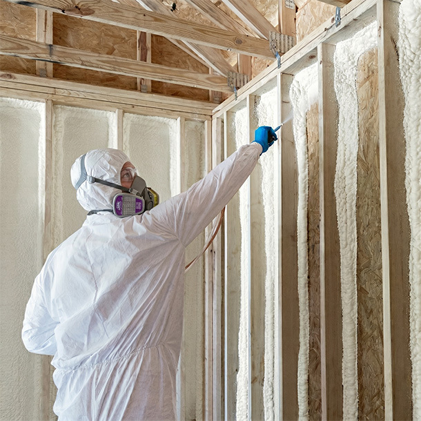 Interior of unfinished wood-framed room with spray foam insulation applied between studs and exposed vaulted ceiling beams; worker exiting in protective suit with foam kits placed on the floor.