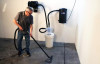 Person cleaning concrete floor in front of garage vacuum and hose reel mounted on wall with a 12 gallon separator bucket sitting on floor between them and attached via vacuum hoses.