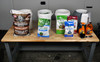 Four buckets sitting on a table with bags of hardwood pellets and various seeds sitting in front of them as well as some cleaning supplies in front of the fourth bucket.