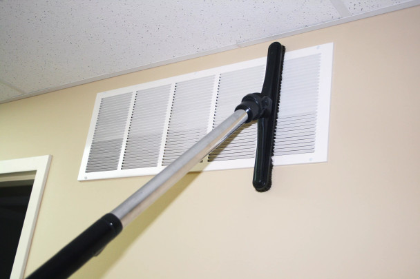 A sidewinder attached to a wand cleaning a vent high up on a wall.