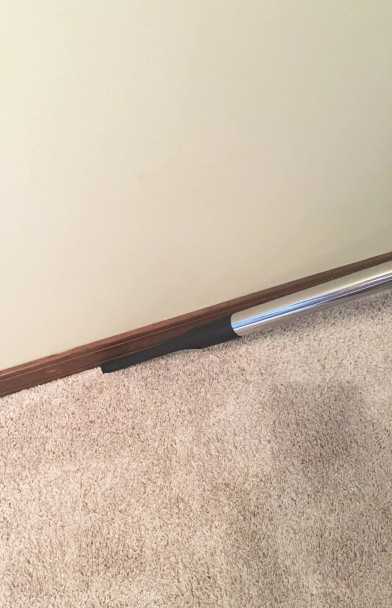 A crevice tool on a wand cleaning the gap between a carpet and a baseboard.