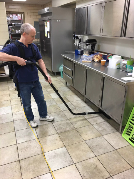 Person cleaning commercial kitchen floor using S-wand and floor brush.