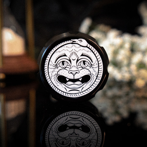 A black unique metal herb grinder with a laughing cat and snake rim around it. the cat has a black crescent moon on its forehead. The bottom side has a crying cat with a snake rim around it.