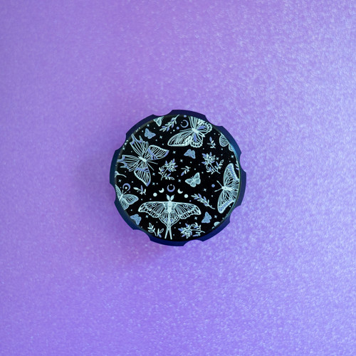 A black metal herb grinder with a mystical print of monarch butterflies, leaves, and crescent moons with purple details.