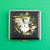 A square case with a whimsical white rabbit that has 3 eyes and is coming out of a gold watch. Around the rabbit are colorful green, red, and orange mushrooms and leaves.This case can fit joints, cigs, and credit cards and birth control pills.