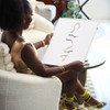 African American woman sitting in chair with green shorts and shoes holding Awujo art print in silver frame on black wall home decor wall art by artist atinuke adeleke for zuri perle