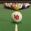 Utah Utes cue ball positioned on green pool table felt in front of a racked set of billiard balls, featuring the red interlocking Utah UU logo with a pool cue lined up for the break.