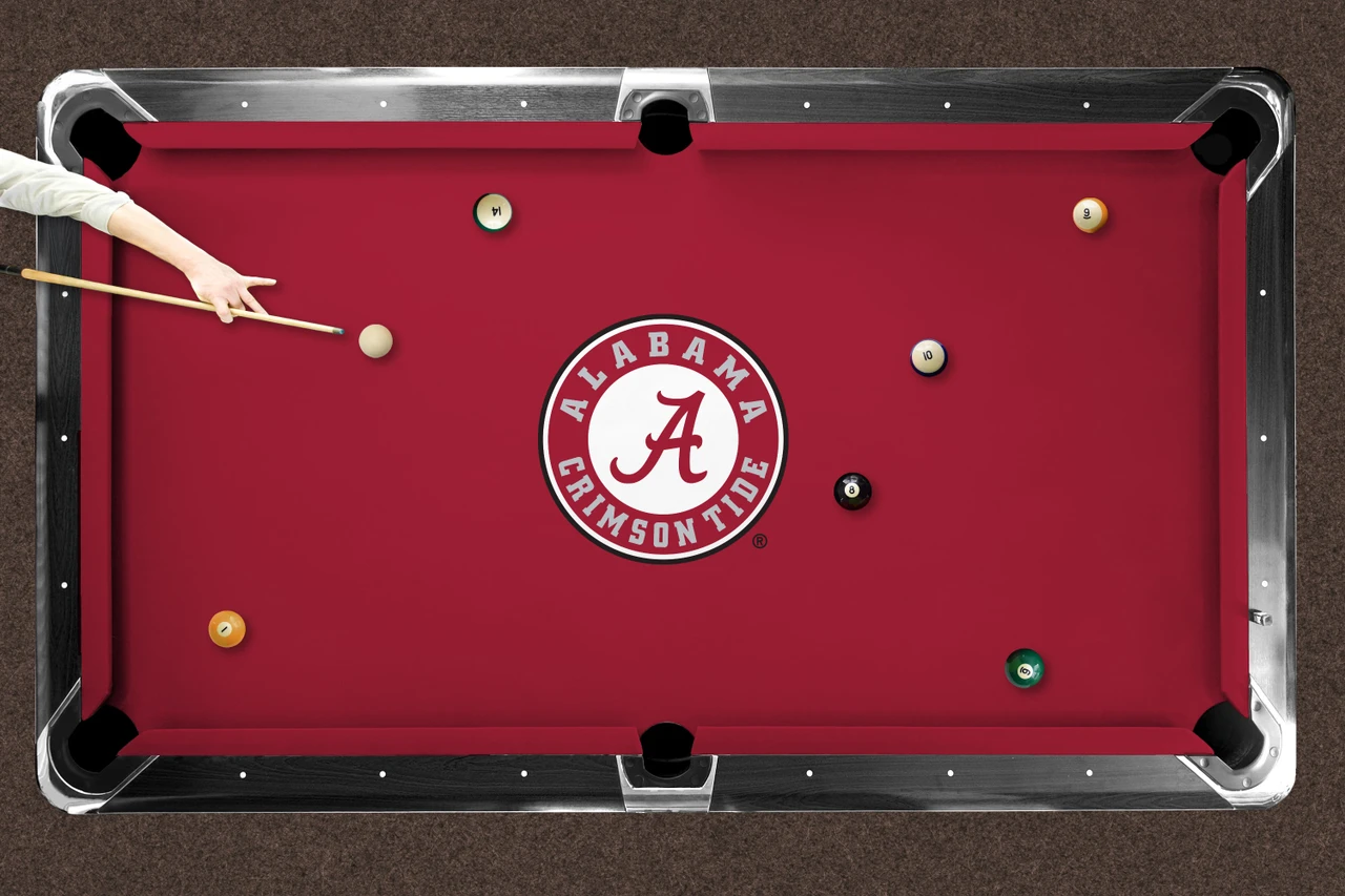 Alabama Crimson Tide logo billiard cloth shown installed on a full-size pool table during play. The crimson playing surface features the large circular Alabama Crimson Tide logo at center with billiard balls positioned across the table and a player preparing to take a shot.
