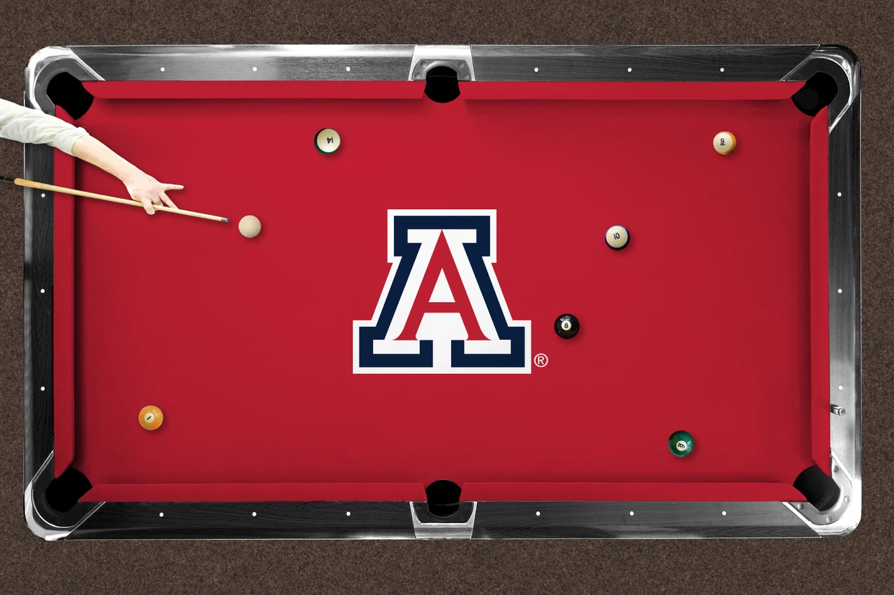 Arizona Wildcats red logo billiard cloth shown installed on a full-size pool table during play. The red playing surface features a large Arizona Wildcats block A logo at center with billiard balls positioned across the table and a player preparing to take a shot.
