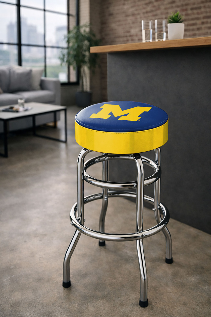 Michigan Wolverines 30" chrome bar stool displayed in a home entertainment setting, featuring a blue and maize team logo seat with a polished chrome base. This college team stool is perfect for adding Michigan spirit to a fan cave, home bar, pool room, or game room.
