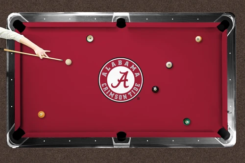Alabama Crimson Tide logo billiard cloth shown installed on a full-size pool table during play. The crimson playing surface features the large circular Alabama Crimson Tide logo at center with billiard balls positioned across the table and a player preparing to take a shot.
