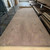 Large walnut veneer sheet with intricate grain patterns lies flat in a spacious warehouse. Stacks of similar sheets are visible on shelves to the right.