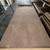 Large walnut veneer sheet with intricate grain patterns lies flat in a spacious warehouse. Stacks of similar sheets are visible on shelves to the right.