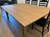 A large wooden veneer table with a smooth surface is surrounded by four black wooden chairs with ladder-back design, set on a light hardwood floor.
