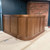 Custom walnut veneer wood-paneled reception counter with a curved base stands against a red brick wall and dark blue door, creating a warm, polished setting.