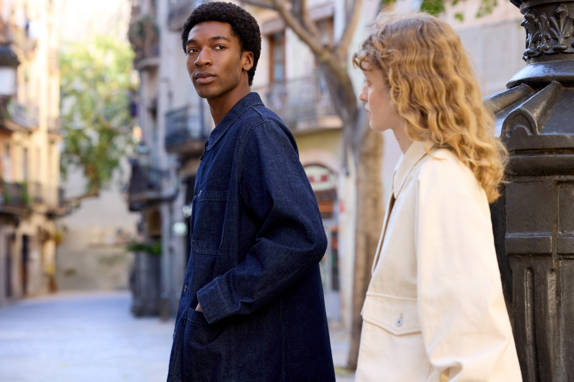 Two people stand on a quiet city street, one in a dark blue MUJI denim jacket and the other in a cream MUJI jacket.
