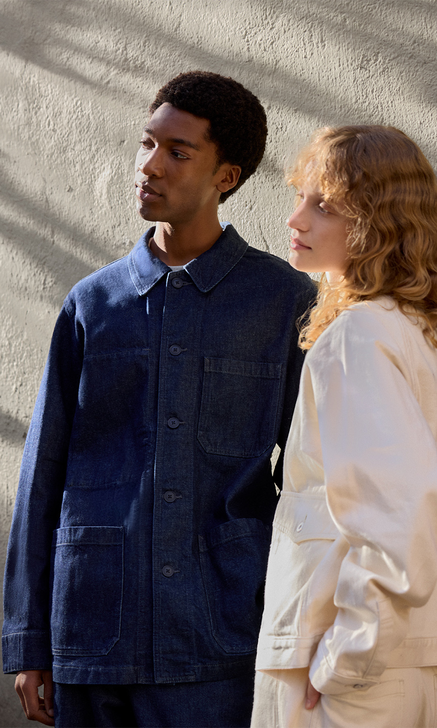 Two people stand on a quiet city street, one in a dark blue MUJI denim jacket and the other in a cream MUJI jacket.