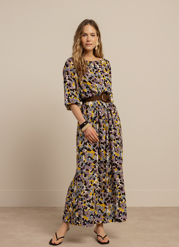 Vrouw staat tegen een neutrale wand en draagt een bruine jumpsuit met korte mouwen en een ceintuur in de taille. De outfit geeft een verfijnde en minimalistische uitstraling met een zachte kleur.