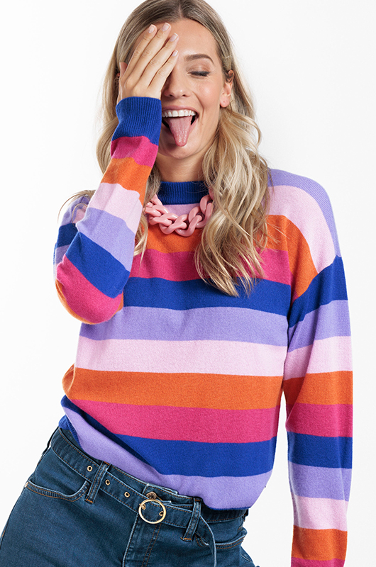 Blond model dat haar tong uitsteekt en een streepjes trui in de kleuren oranje, roze, blauw, paars en oudroze draagt