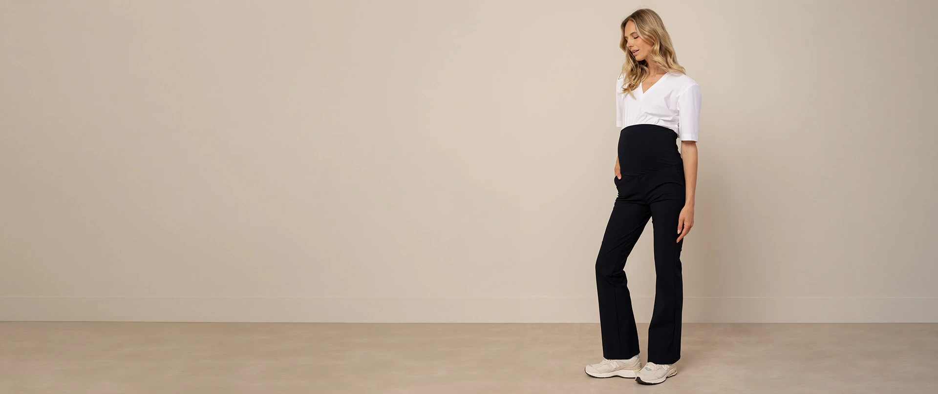 Zwangere vrouw draagt een wit T-shirt met een donkerblauwe zwangerschapsbroek en houdt haar buik vast terwijl ze licht glimlacht. De outfit laat zien hoe maternity kleding comfort en stijl combineert in een minimalistische look.