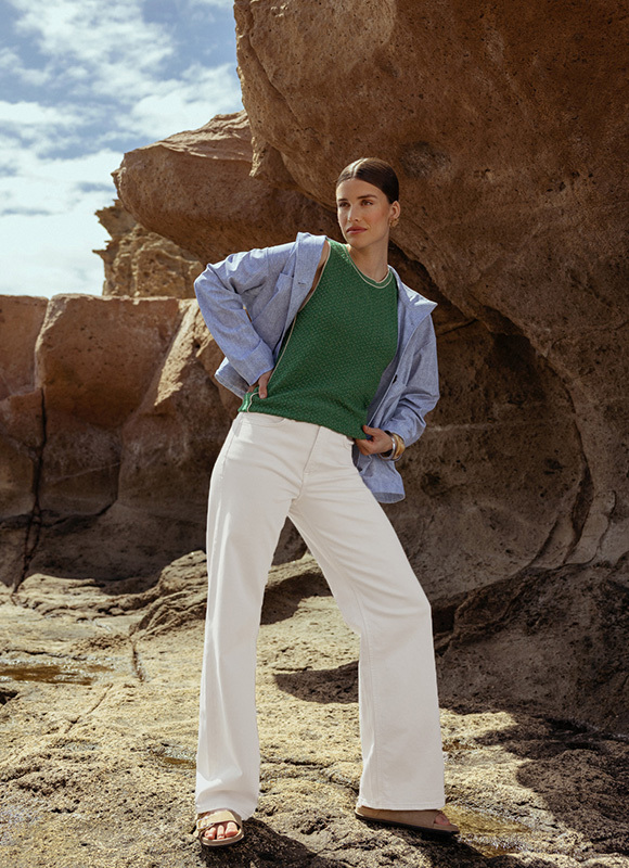 Vrouw poseert zelfverzekerd tussen grote rotsen aan de kust en draagt een groene top met een lichtblauwe blouse en wijde witte broek. Ze kijkt opzij terwijl het zonlicht de natuurlijke stenen en rustige strandsfeer benadrukt.