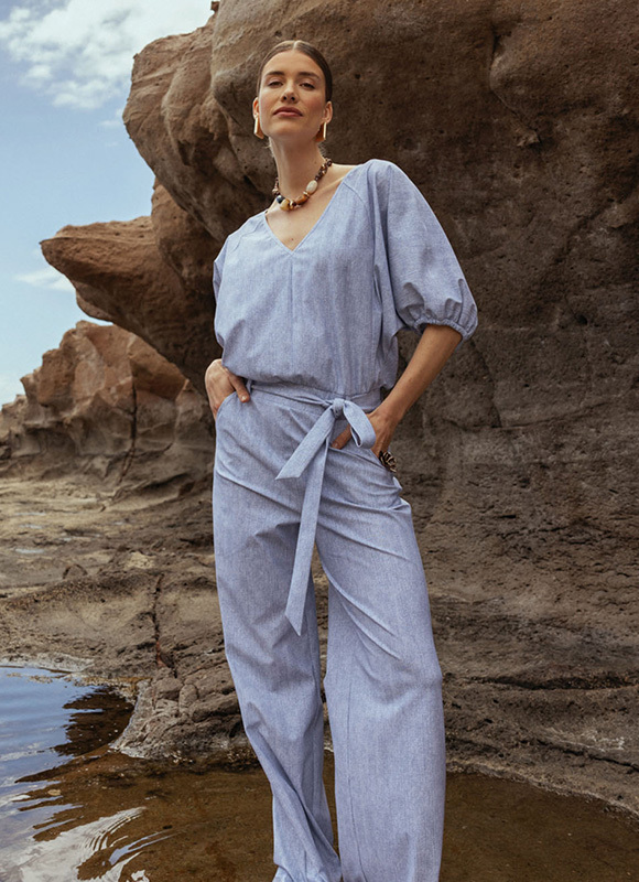 Vrouw in een lichtblauwe jumpsuit staat ontspannen tussen rotsen aan de kust met haar handen in de zakken en kijkt zelfverzekerd vooruit. Ze draagt opvallende sieraden terwijl het natuurlijke landschap en een kleine waterplas de rustige, zomerse setting benadrukken.